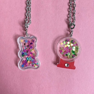 May include: Two necklaces with clear plastic charms filled with colorful glitter. One charm is a bear shape and the other is a gumball machine shape.