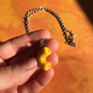 May include: A yellow macaroni-shaped pendant on a silver chain necklace. The pendant is a realistic depiction of a macaroni noodle.