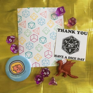 May include: A thank you card with a dice pattern and the text "Thank You Have A Dice Day". The card is surrounded by purple dice and a small red dragon figurine.