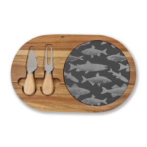 Puede incluir: Tabla de queso ovalada de madera con una zona de servicio gris oscuro estampada con peces. Incluye un cuchillo y un tenedor para queso con mangos de madera. La tabla es de madera marrón claro.
