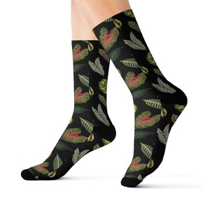 Puede incluir: Calcetines negros con un patrón repetido de hojas verdes, rojas y grises. Las hojas varían en forma y tamaño, creando un diseño botánico. Los calcetines se llevan en las piernas de una persona.