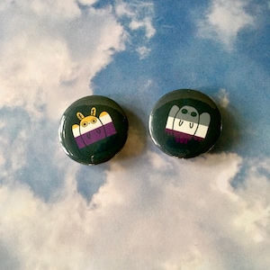 Puede incluir: Dos pines redondos con fondo negro y contornos blancos. El pin de la izquierda presenta un animal de dibujos animados con rayas amarillas, blancas y moradas. El pin de la derecha presenta un fantasma de dibujos animados con rayas blancas, moradas y negras.