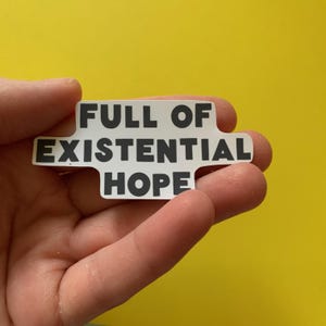 May include: A white sticker with the text "FULL OF EXISTENTIAL HOPE" in black, held against a yellow background. The sticker has a unique shape with cut-out sections. The text is in a bold, sans-serif font.