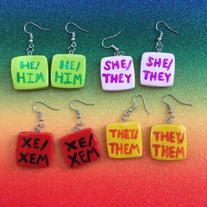 May include: Six pairs of square-shaped earrings with different pronoun combinations. The earrings are made of clay and have a glossy finish. The pronouns are written in black, red, and pink paint. The earrings are on a rainbow background.