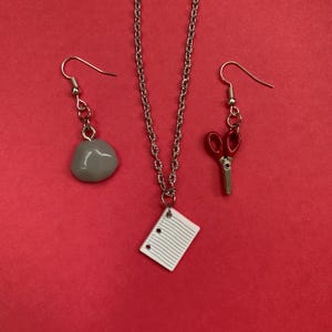May include: A jewelry set featuring a necklace and two earrings. The necklace has a silver chain with a pendant of a small, white notepad. One earring is a gray stone, and the other is a pair of red scissors.