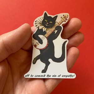 May include: A sticker featuring a black cat playing a banjo, wearing a ruffled collar. The cat has yellow eyes and is standing on its hind legs. The text "off to commit the sin of empathy!" is printed at the bottom.