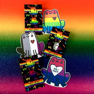 May include: Set of six enamel pins featuring frogs with rainbow colors and pride flag designs. The pins have the text "World's Queerest Pride Frog Pins" and "He/They".