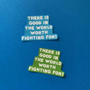 May include: Two stickers with the text "THERE IS GOOD IN THE WORLD WORTH FIGHTING FOR!". One sticker is light blue with white text, and the other is green with white text. The stickers are on a blue background.