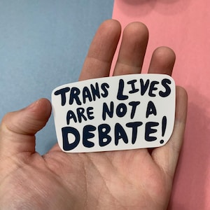 May include: A rectangular white sticker with rounded corners featuring the text TRANS LIVES ARE NOT A DEBATE! written in a bold, dark blue, hand-lettered font. The sticker has a smooth finish and is presented against a neutral background.