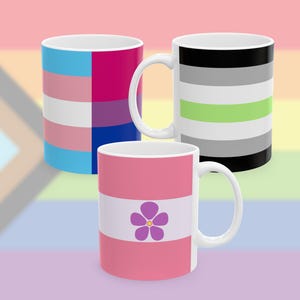 Puede incluir: Tres tazas de cerámica blanca con diseños coloridos. Una taza presenta la bandera del orgullo transgénero, otra la bandera del orgullo arromántico, y la tercera tiene un diseño rosa y blanco con una flor morada. Las tazas tienen asas blancas.
