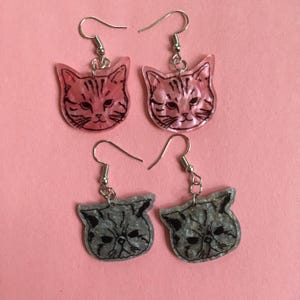 May include: A pair of pink cat face earrings and a pair of gray cat face earrings. The cat faces are outlined in black and have a glitter finish.