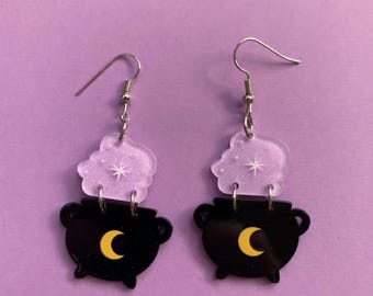 May include: A pair of dangle earrings featuring a black cauldron design with a yellow crescent moon and a translucent cloud with a star. The earrings have silver-colored hooks and are set against a purple background.