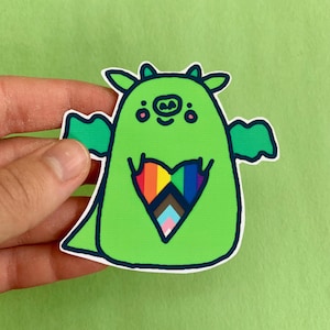 May include: A green sticker featuring a cartoon monster with a heart-shaped design. The heart is filled with a rainbow and pride flag colors. The monster has small horns, wings, and a smiling face. The sticker is outlined in dark green.