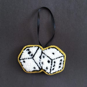 May include: A white dice ornament with black dots, a gold glitter border, and a black ribbon for hanging. The dice are made of a textured material, and the ornament is set against a dark background. A fun, decorative item.