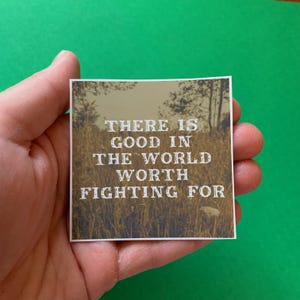 May include: Square sticker with the text "THERE IS GOOD IN THE WORLD WORTH FIGHTING FOR" in white, set against a sepia-toned background of a field and trees. The sticker is held in a hand against a green background.