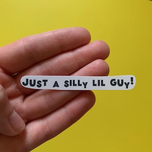 May include: A white sticker with the text "JUST A SILLY LIL GUY!" in black, held against a yellow background. The sticker is rectangular with rounded edges.