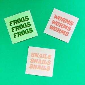 May include: Three colorful square stickers with the words "Frogs", "Worms", and "Snails" printed in bold, stacked letters. The "Frogs" sticker is green with dark green letters, the "Worms" sticker is pink with light pink letters, and the "Snails" sticker is light pink with dark pink letters.