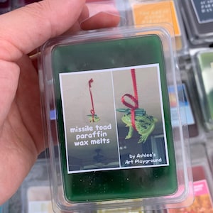 May include: A green wax melt in a clear plastic container. The label reads "missile toad paraffin wax melts" with a cartoon frog illustration. The product is from "Ashlee's Art Playground."