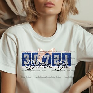 Puede incluir: Una camiseta blanca de manga corta con un diseño gráfico que muestra los números 30720 en purpurina azul, un lazo rosa claro sobre los números y el texto Dalton, Ga en letra cursiva negra. El diseño incluye pequeños detalles perlados.
