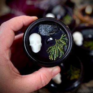 May include: A black cauldron-shaped candle holder with a white skull, a small grey stone, and a sprig of green foliage. The candle holder is sitting on a table with other similar candle holders.