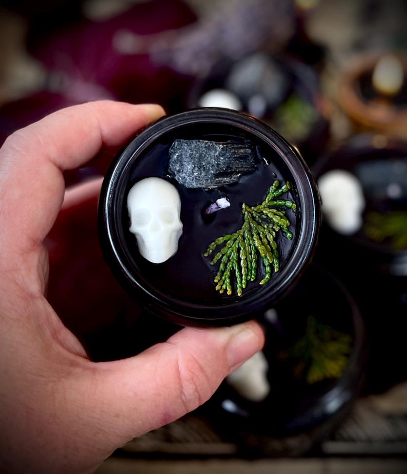 May include: A black cauldron-shaped candle holder with a white skull, a small grey stone, and a sprig of green foliage. The candle holder is sitting on a table with other similar candle holders.
