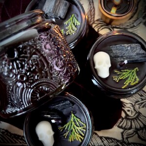 May include: Four black glass candle jars with lids. Each jar contains a black candle with a skull, a piece of black charcoal, and green sprigs. The jars are on a patterned surface.