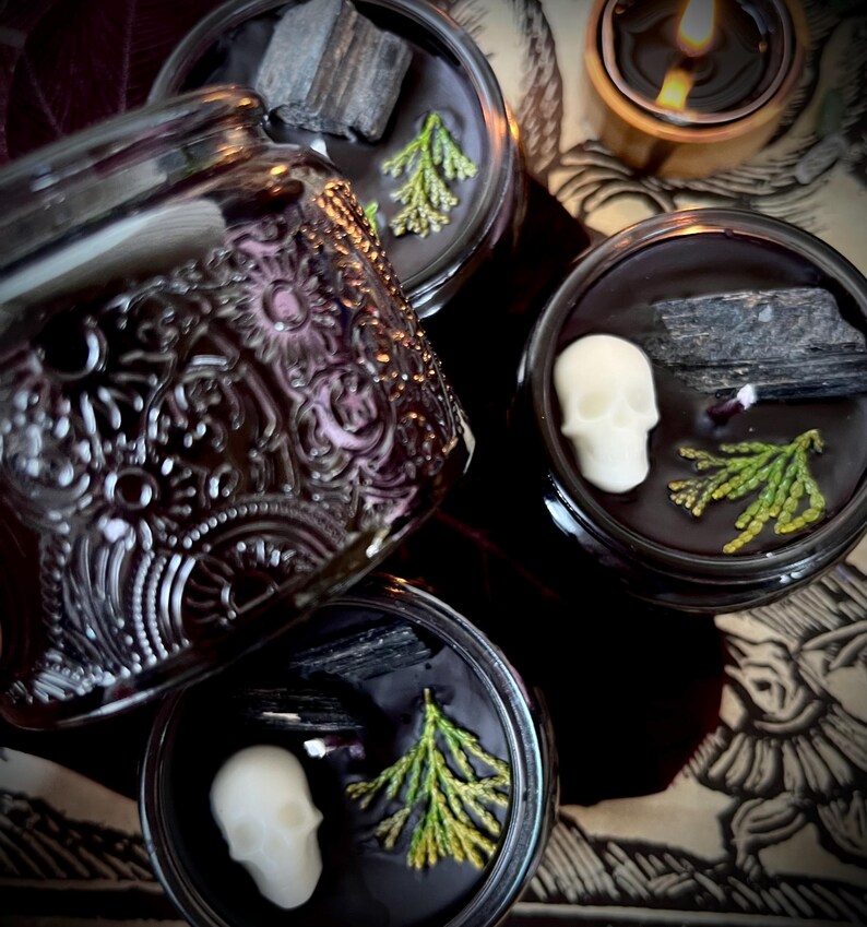 May include: Four black glass candle jars with lids. Each jar contains a black candle with a skull, a piece of black charcoal, and green sprigs. The jars are on a patterned surface.