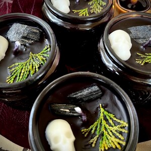 May include: Four black glass candle jars with lids. Each jar contains a black candle with a white skull, a piece of black charcoal, and a sprig of green foliage.