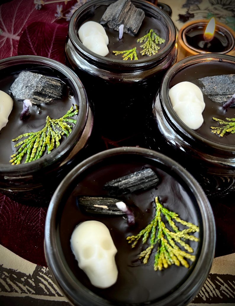 May include: Four black glass candle jars with lids. Each jar contains a black candle with a white skull, a piece of black charcoal, and a sprig of green foliage.