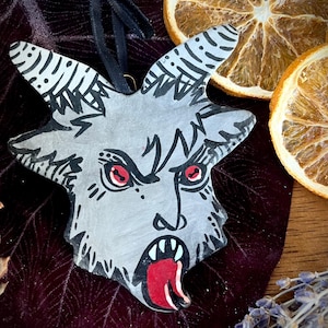 Op de afbeelding: Een grijs, stervormig ornament met een duivels gezicht, rode ogen en een open mond met een rode tong. Het ornament heeft zwarte accenten en een zwart lint om op te hangen. Gedroogde sinaasappelschijfjes en lavendel in de achtergrond.
