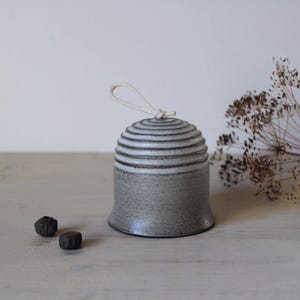 Könnte beinhalten: Ein glockenförmiges Keramikobjekt mit einer gesprenkelten grauen Basis und einem weiß glasierten, gerippten Oberteil. Eine weiße Kordelschlaufe ist oben mittig befestigt. Links neben dem Keramikstück liegen zwei kleine, dunkle, strukturierte Perlen.