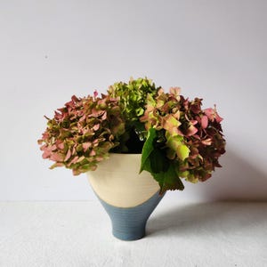 Può includere: Un vaso in ceramica bicolore, color crema nella parte superiore e blu nella base, contenente un bouquet di ortensie. I fiori sono un mix di verde e rosa, con foglie verdi. Il vaso è su una superficie bianca.
