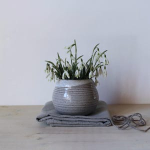 Peut inclure: Un petit vase rond en céramique grise avec des rainures horizontales, rempli de fleurs de perce-neige blanches. Le vase est posé sur un tissu en lin gris plié. Un paquet de ficelle grise repose à droite, suggérant un thème rustique et naturel.