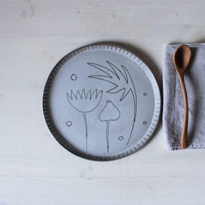 Può includere: Un piatto rotondo in ceramica grigia con un disegno floreale disegnato a mano in nero. Il piatto ha un bordo strutturato ed è accompagnato da un cucchiaio di legno e un tovagliolo di lino grigio. Il design del piatto presenta tre fiori stilizzati e piccoli cerchi.