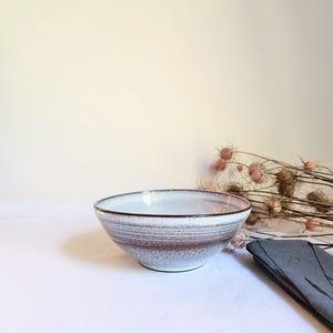 Può includere: Una ciotola in ceramica con interno smaltato bianco ed esterno maculato con bande orizzontali marroni e grigie. La ciotola ha una forma arrotondata con un bordo marrone scuro e una piccola base, caratterizzata da una finitura testurizzata.