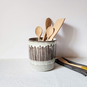 Può includere: Un portautensili in ceramica con un design a righe bianche e marroni, pieno di cucchiai di legno e una spatola. Il portautensili è cilindrico e poggia su una superficie bianca. Una forchetta di legno è appoggiata su un tovagliolo grigio scuro.