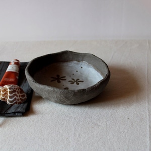 Può includere: Una ciotola in ceramica grigio scuro fatta a mano con un interno chiaro con due disegni floreali marroni. La ciotola ha un esterno ruvido e strutturato. Un tubetto di crema per le mani rossa e una conchiglia sono visibili.
