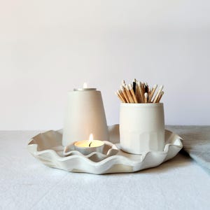 Pode incluir: Um conjunto de cerâmica branca composto por uma bandeja grande com bordas onduladas, um castiçal alto com uma vela acesa, um prato pequeno a condizer com uma vela de chá acesa e um recipiente cilíndrico cheio de fósforos de madeira.