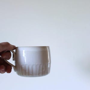 Peut inclure: Une tasse en céramique blanche avec une anse, tenue par une main. La tasse présente des détails verticaux côtelés sur la partie inférieure et une finition lisse et brillante. La tasse est conçue pour les boissons chaudes.