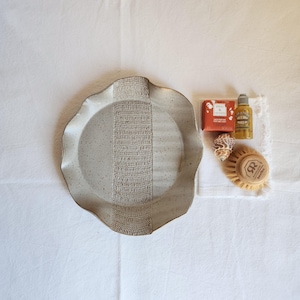 May include: A speckled, off-white ceramic plate with a textured, woven-like design down the center. Accompanying items include a bar of soap, a small bottle, a seashell, and a wooden dish brush.