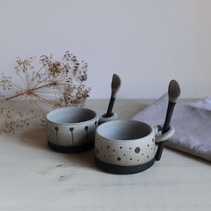 Puede incluir: Dos tazas de cerámica con cucharas a juego. Las tazas son de color gris claro con bases negras y patrones de puntos. Las cucharas tienen mangos negros y cabezas de cuchara grises. Adecuado para café o té.