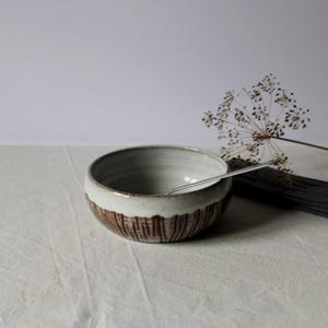 Può includere: Una ciotola in ceramica con interno smaltato bianco ed esterno marrone testurizzato con linee verticali intagliate. Un cucchiaio in metallo argentato riposa all'interno della ciotola e un gambo di pianta essiccata si trova accanto.