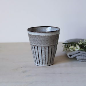 Könnte beinhalten: Ein handgefertigter Keramikbecher mit einem strukturierten Design mit vertikalen Linien. Der Becher hat ein Grau-Weiß-Farbschema mit einem dunkelbraunen Rand. Der Becher ist neutral und eignet sich für heiße oder kalte Getränke.