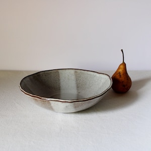 Può includere: Una ciotola in ceramica fatta a mano con finitura grigio chiaro macchiettata e bordo marrone scuro. La ciotola ha una forma irregolare e organica. Una pera matura con buccia marrone e lungo gambo è accanto alla ciotola.