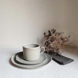 May include: A neutral-toned ceramic mug and two plates with a textured finish. The plates have a decorative edge. A folded gray napkin and dried flowers are arranged nearby on a white surface. The tableware is suitable for a minimalist aesthetic.