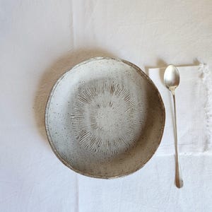 Può includere: Una ciotola in ceramica fatta a mano, macchiata, con un design radiale strutturato al centro. La ciotola è bianco sporco con un bordo leggermente ruvido. Un lungo cucchiaio d'argento è appoggiato accanto su un tovagliolo di lino bianco.