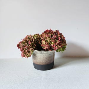 Może przedstawiać: Biały ceramiczny kubek z czarnym uchwytem i napisem "But First, Coffee" czarną, pogrubioną czcionką. Kubek stoi na białej powierzchni. Kubek ma około 10 cm wysokości.