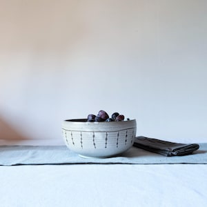 Könnte beinhalten: Eine Keramikschale gefüllt mit dunklen Trauben, auf einem hellblauen Leinenläufer. Die cremefarbene Schale hat einen dunklen Rand und ein sich wiederholendes vertikales Punktmuster. Eine gefaltete dunkelgraue Serviette befindet sich rechts.