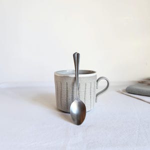 Può includere: Un cucchiaio in acciaio inossidabile è appoggiato davanti a una tazza di ceramica grigia con linee nere verticali. La tazza ha un manico e poggia su una superficie bianca. Il cucchiaio è lungo circa 18 cm.