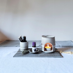 May include: A ceramic oil burner with a lit tea light candle, a small ceramic cup containing black matches, a small bottle of essential oil with a purple cap, and a sprig of dried lavender, all arranged on a rectangular ceramic tray.